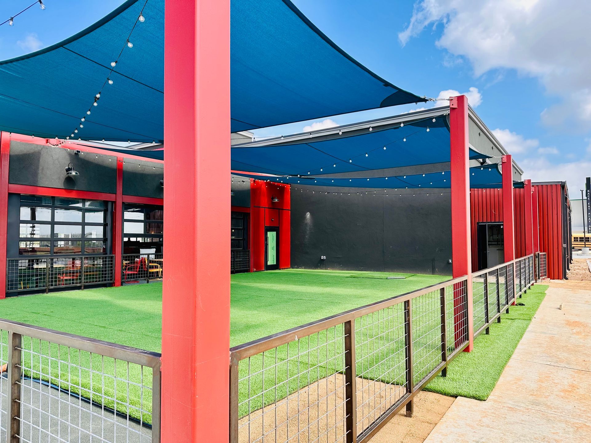 Outdoor patio with blue shade sails, red columns, green turf, and a black wall fence