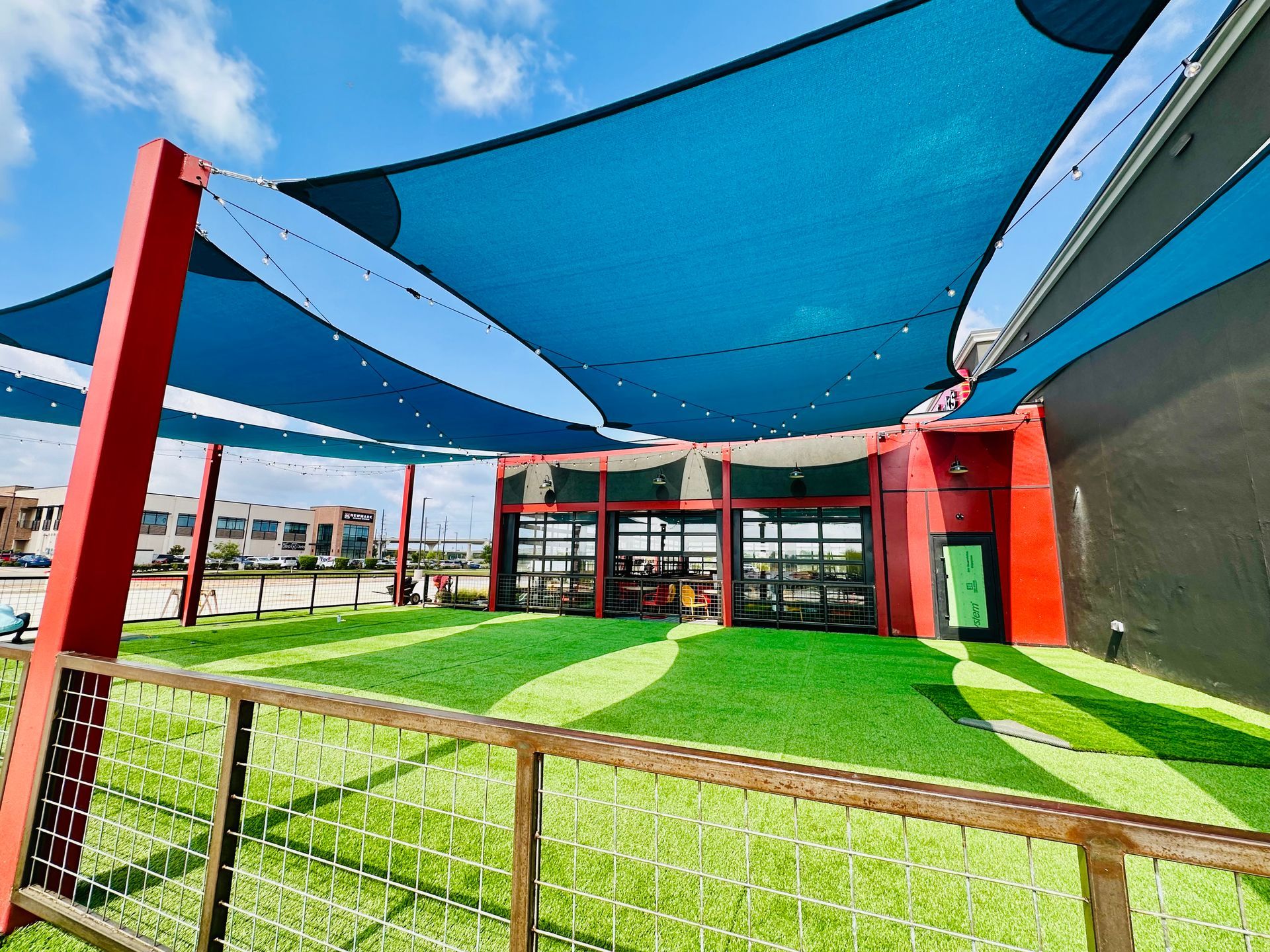 Modern outdoor courtyard with blue shade sails, red columns, green grass, and a building backdrop