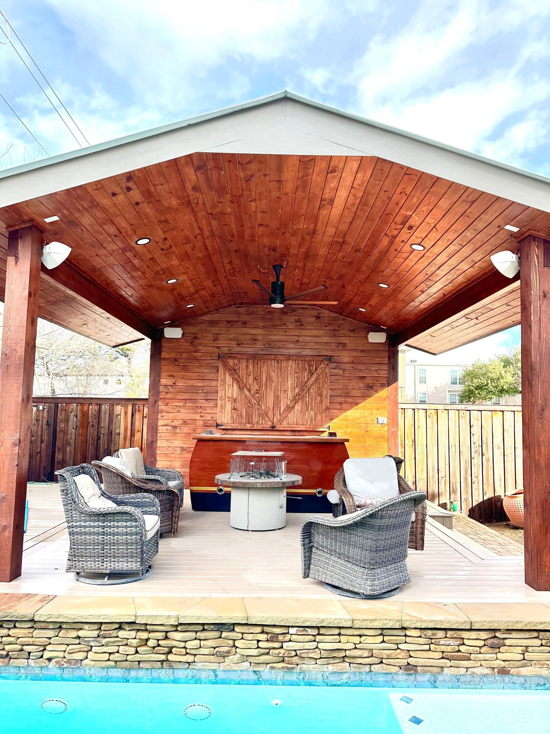 Covered patio with wood ceiling, wicker chairs, and a small table beside a pool and fence.