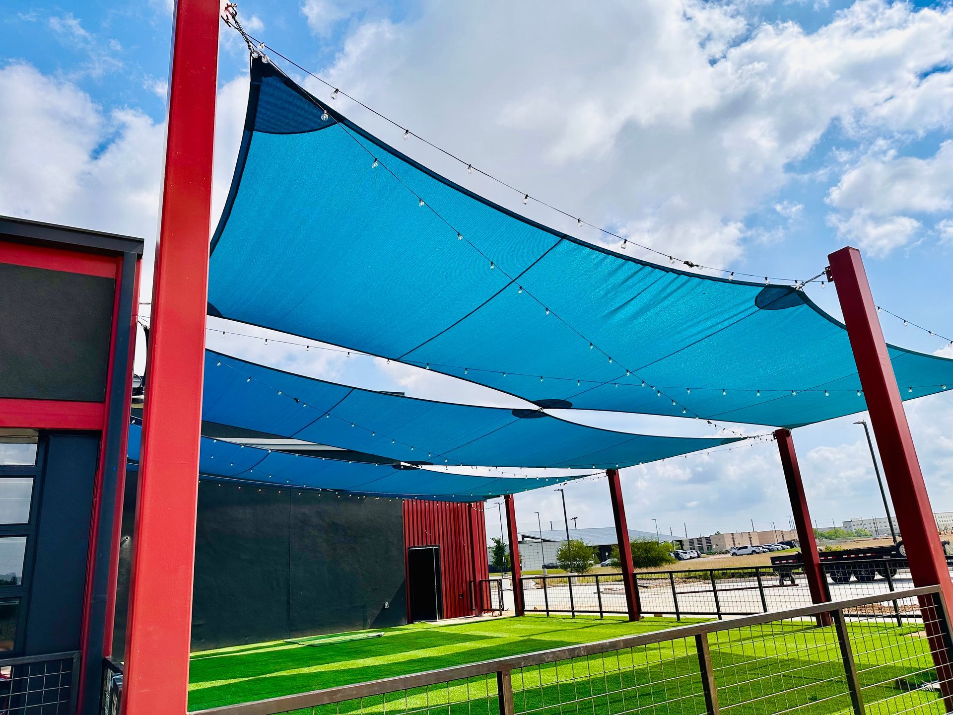 Blue shade sails over a grassy outdoor area with red poles and a cloudy sky.