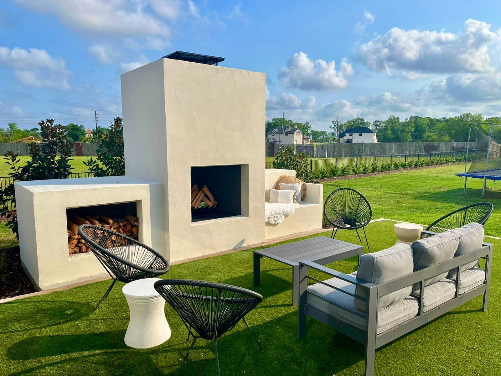 Modern outdoor patio with white fireplace, black chairs, sofa, table, and green lawn under a blue sky