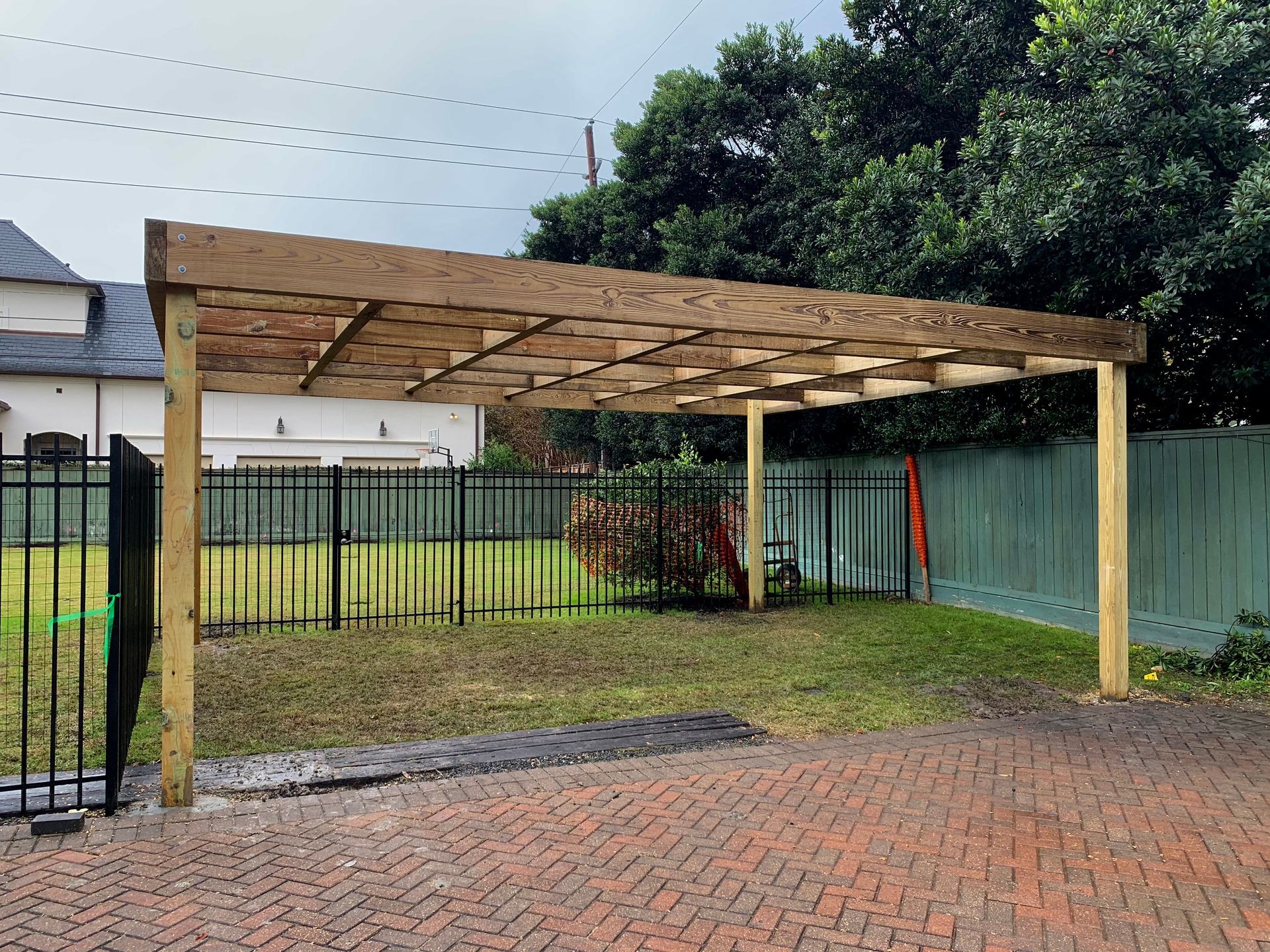 Wooden carport beside a brick driveway and fenced yard under a cloudy sky