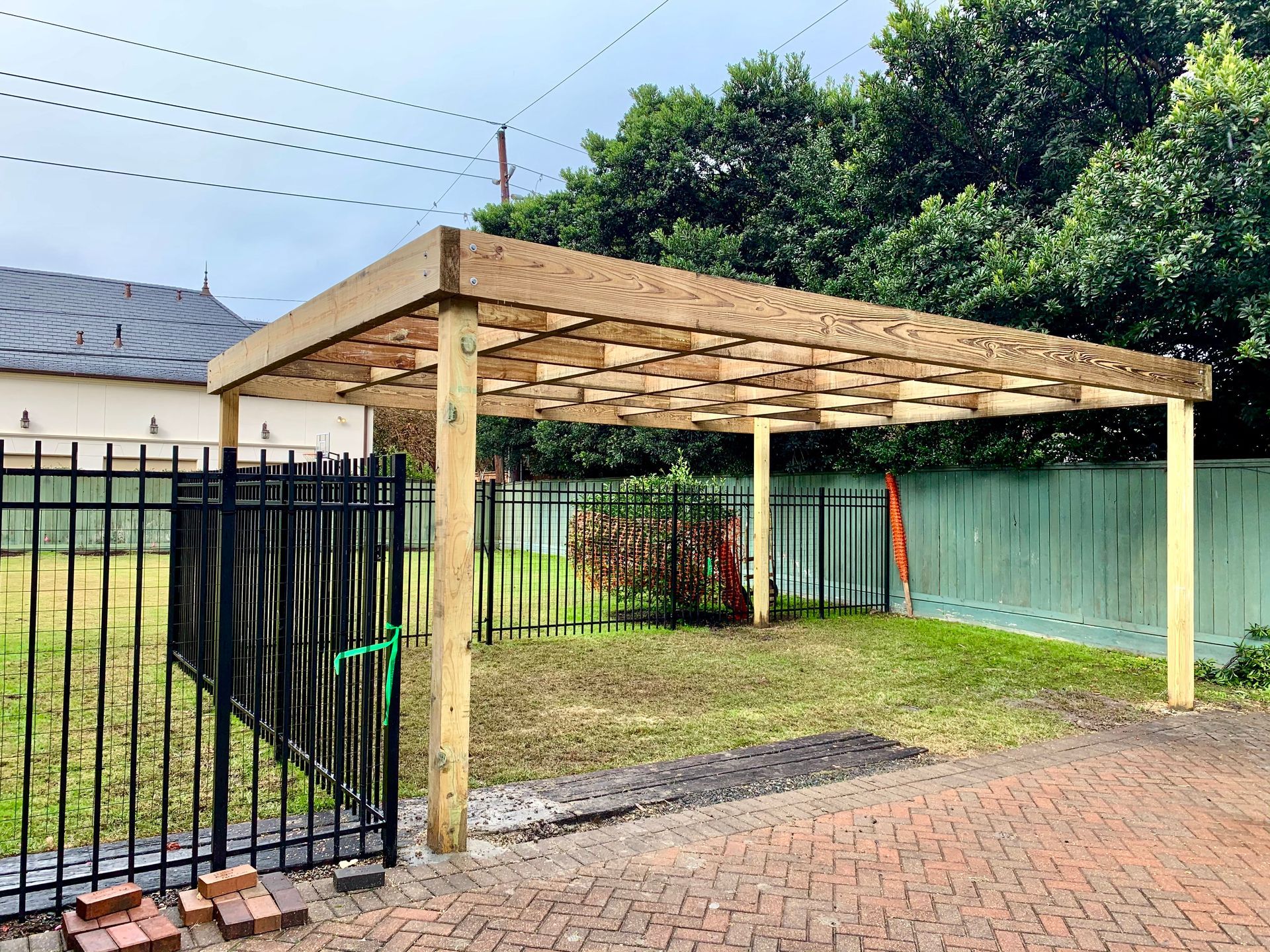 Covered carport with wooden frame beside a black fence and paved driveway in a residential yard