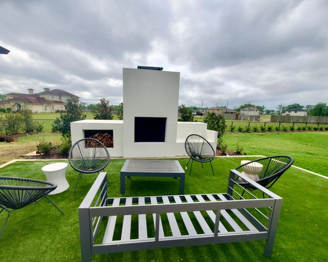 Outdoor patio with gray metal seating around a fire pit, facing a white fireplace on a green lawn.
