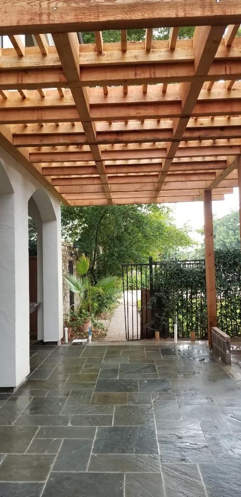 Covered stone patio with wooden pergola, white columns, and steps leading to a garden.