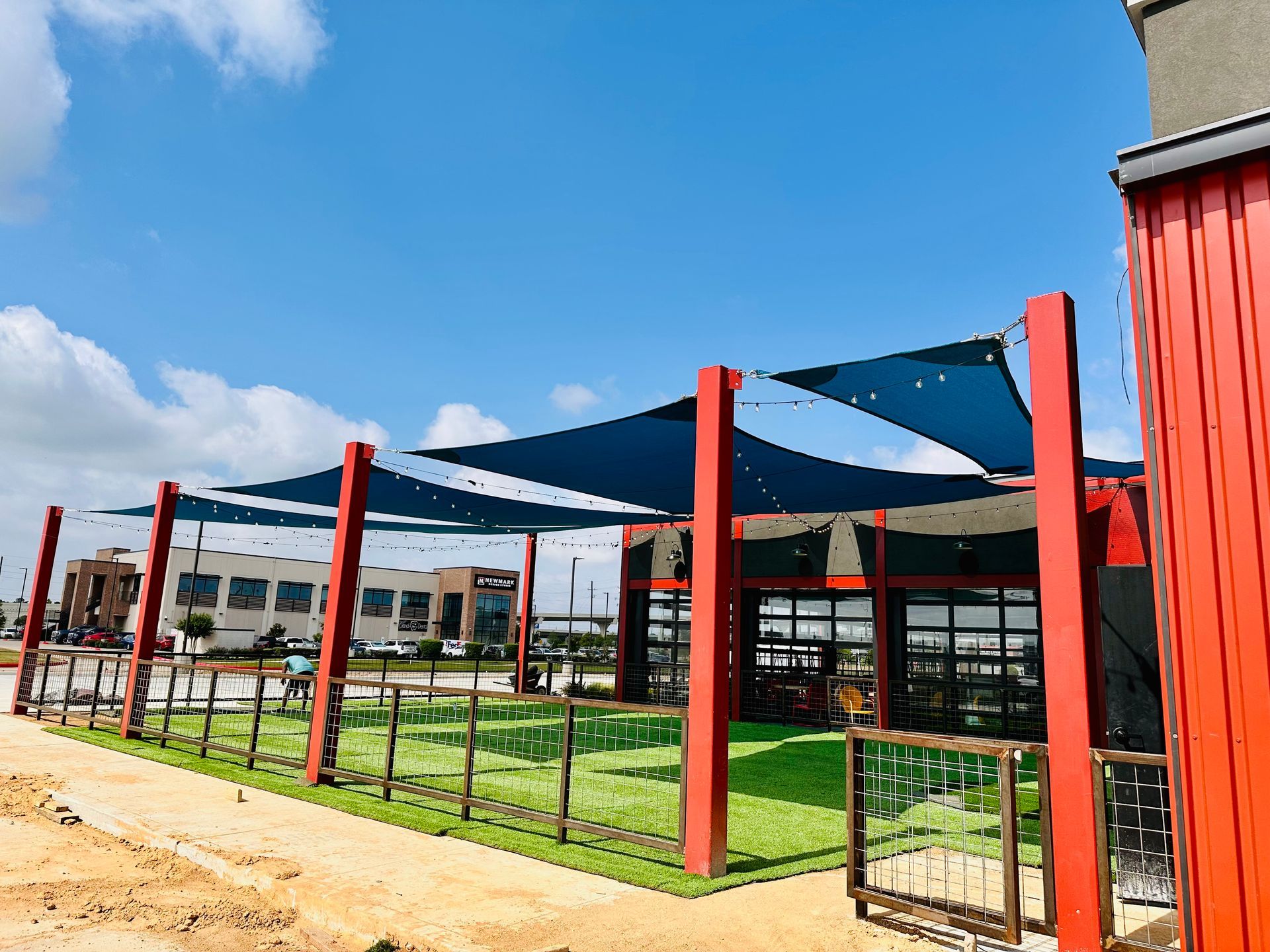 Outdoor café seating with red posts, blue shade sails, and a grassy patio under a bright sky