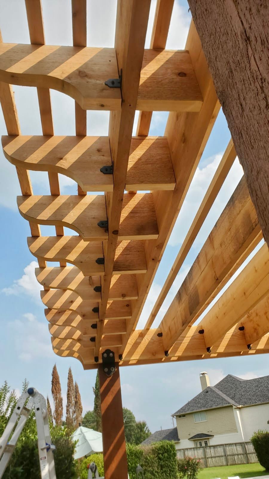Wooden pergola beams overhead in a sunny backyard with houses and trees in the background
