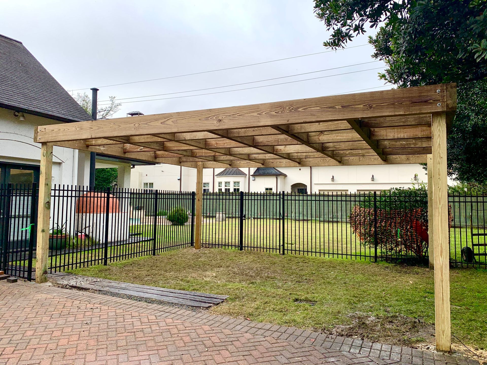 Wooden pergola over a grassy yard with a black fence and brick paving beside houses