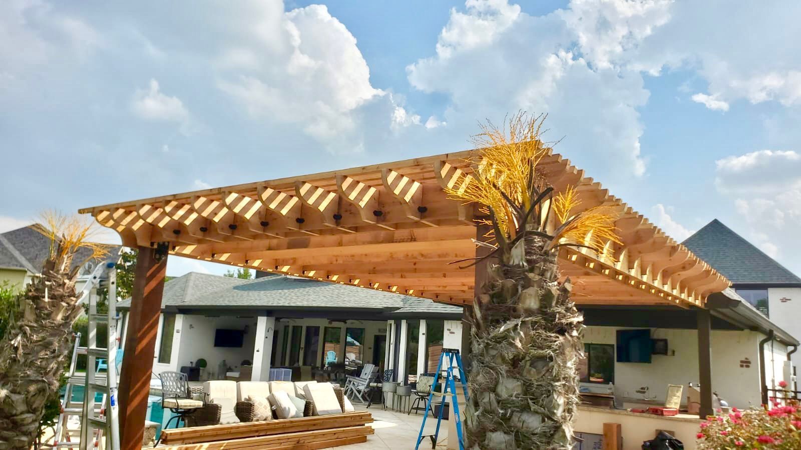 Outdoor patio with a wooden pergola, stone columns, and seating in front of a house under a blue sky