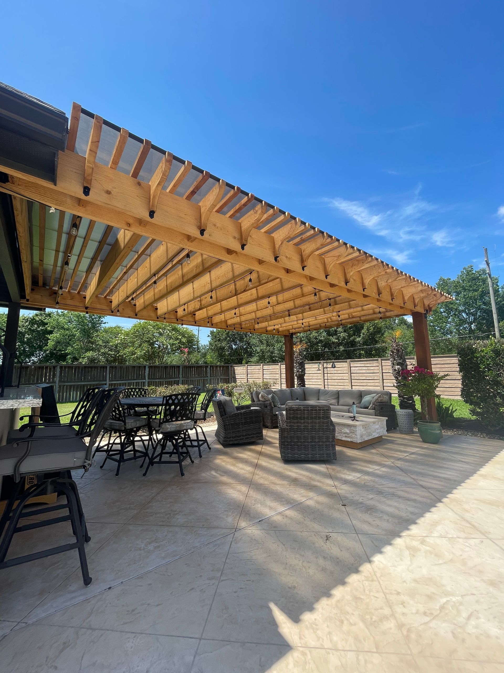 Outdoor patio with wooden pergola, tables, chairs, and stone fire pit under a blue sky