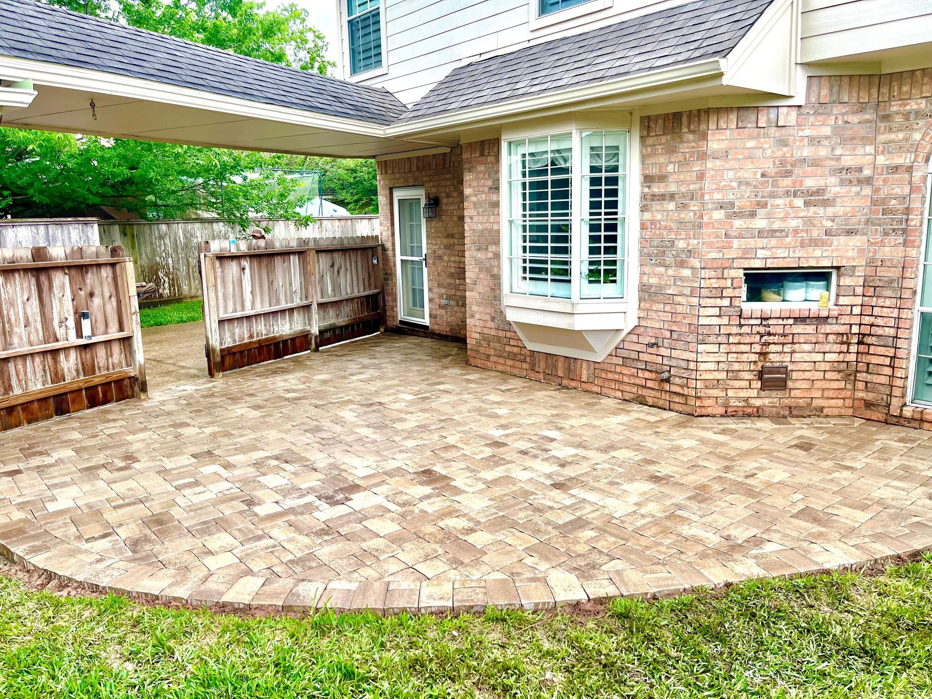 Backyard patio with brick pavers, brick house wall, white bay window, and wooden fence gates