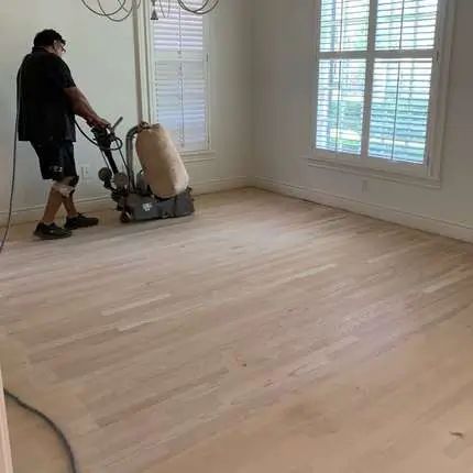 Worker using a floor sander on light wood flooring in a bright room with white shutters.