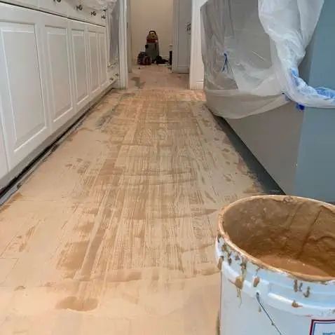 Paint bucket on a kitchen floor during wood floor refinishing, with cabinets and plastic-covered furniture nearby