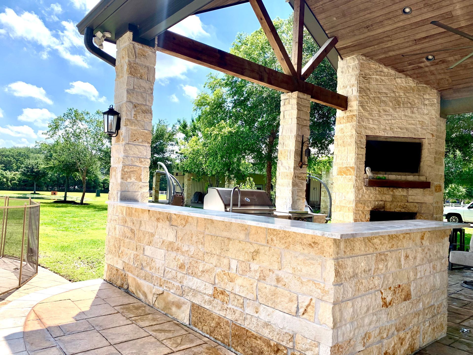 Covered outdoor stone grill and bar under a wooden pergola on a sunny patio by a grassy lawn