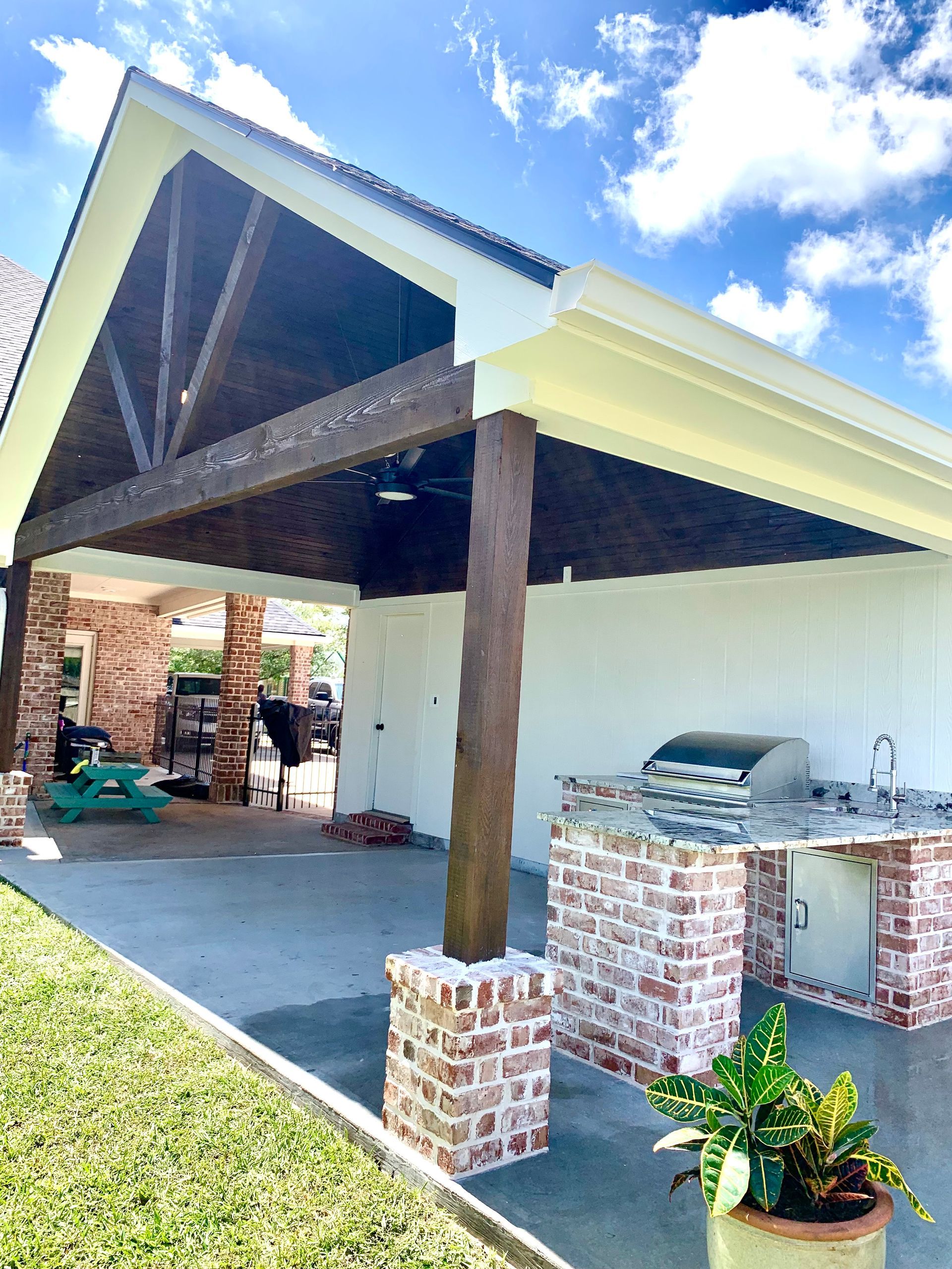 Carport with exposed wood beams, brick columns, and a grill beside a white house under a blue sky.