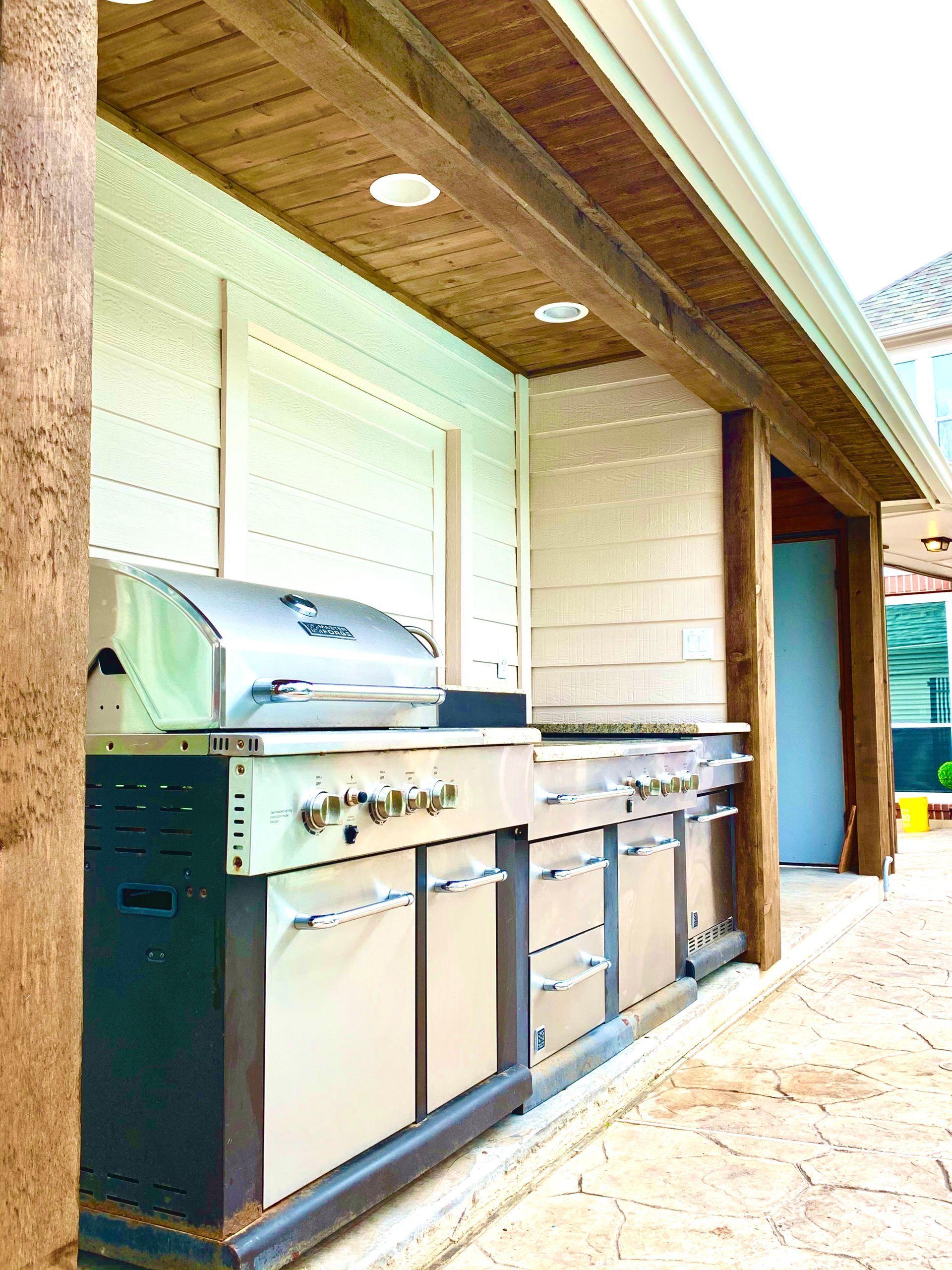 Outdoor kitchen with stainless steel grill and cabinets under a wooden patio roof