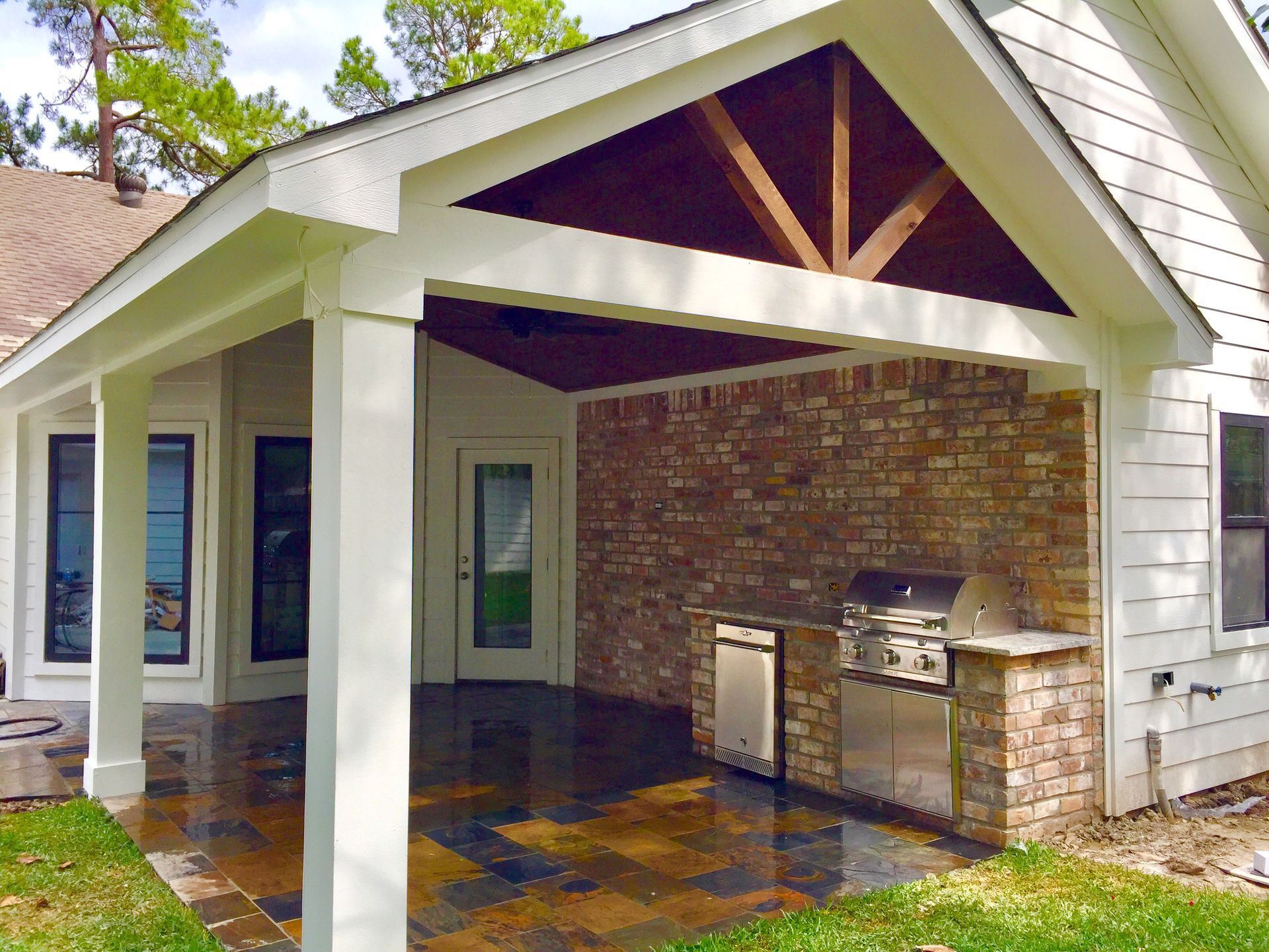 Covered patio with brick fireplace, built-in grill, and tiled floor under a white gabled roof