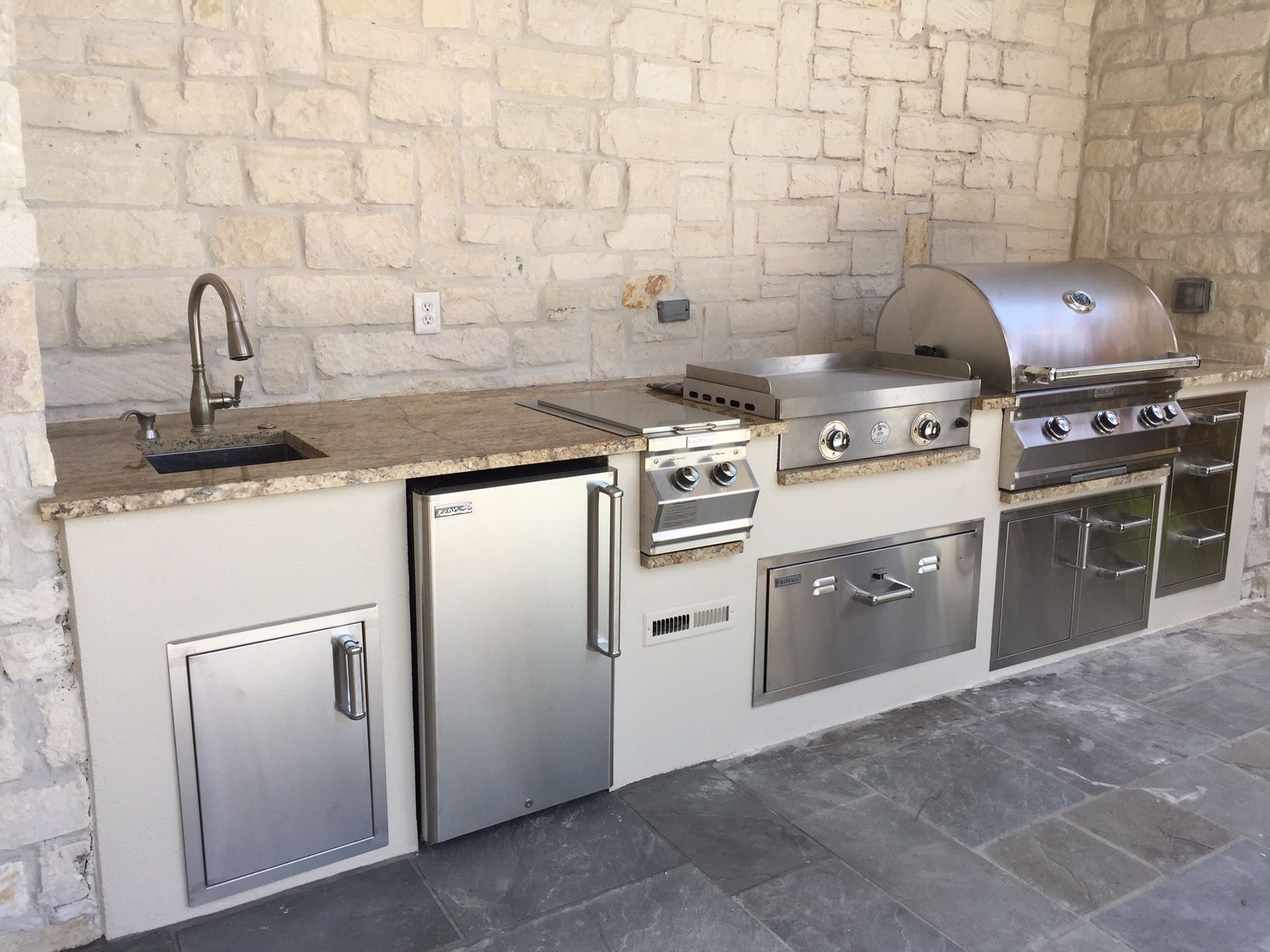 Outdoor built-in stainless steel grill, sink, and storage cabinets against a stone wall.