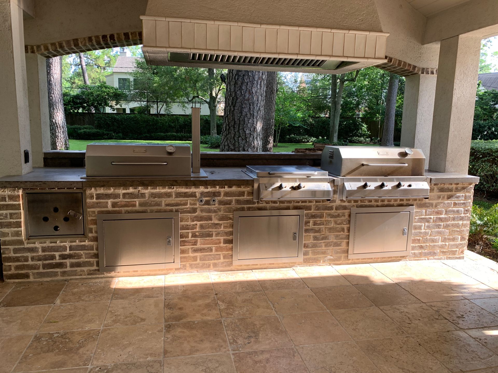 Covered outdoor kitchen with brick counter, built-in grill, sink, cabinets, and stone floor.