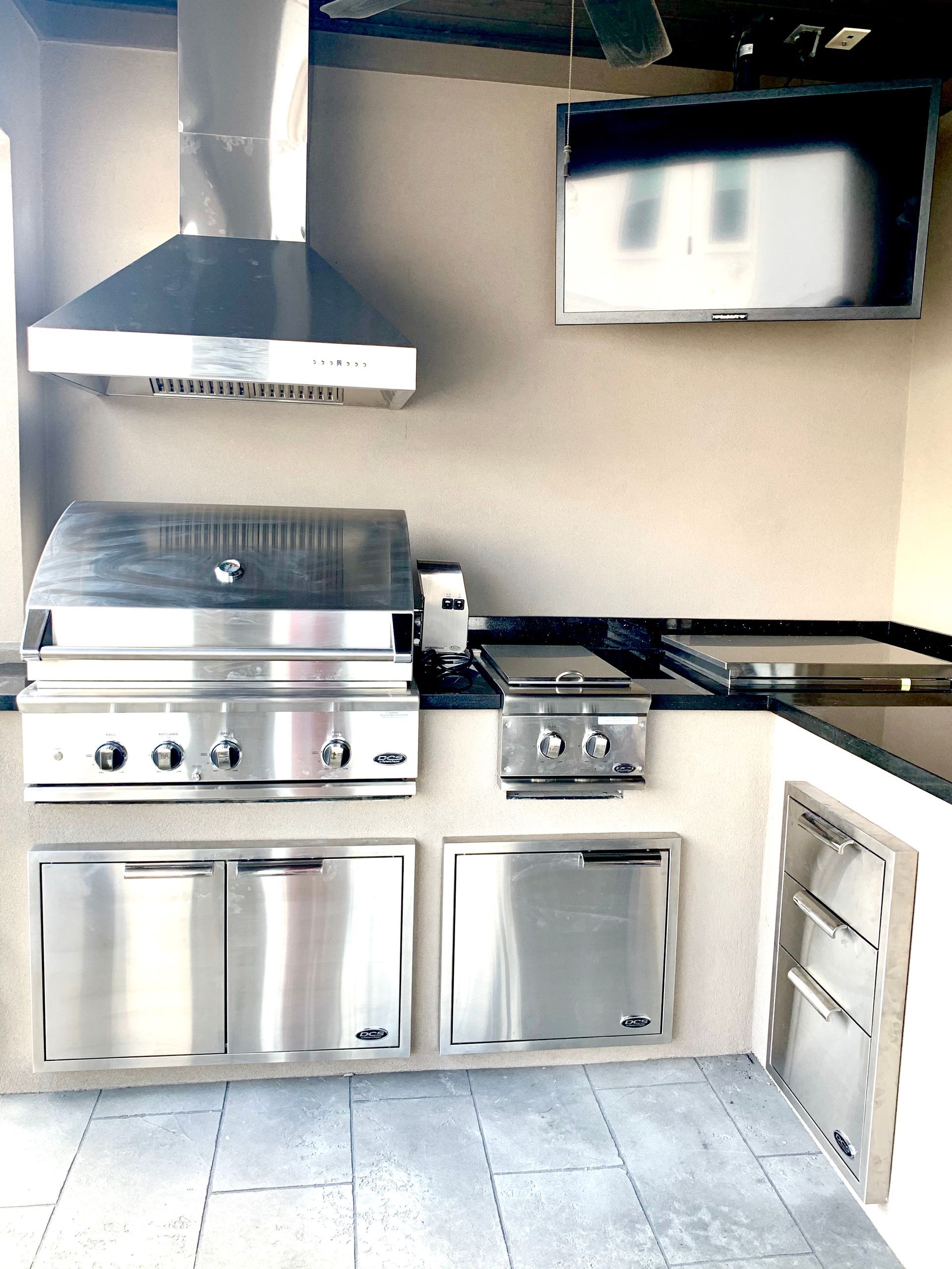 Stainless steel outdoor kitchen with grill, hood, cabinets, and side burner on a tiled patio