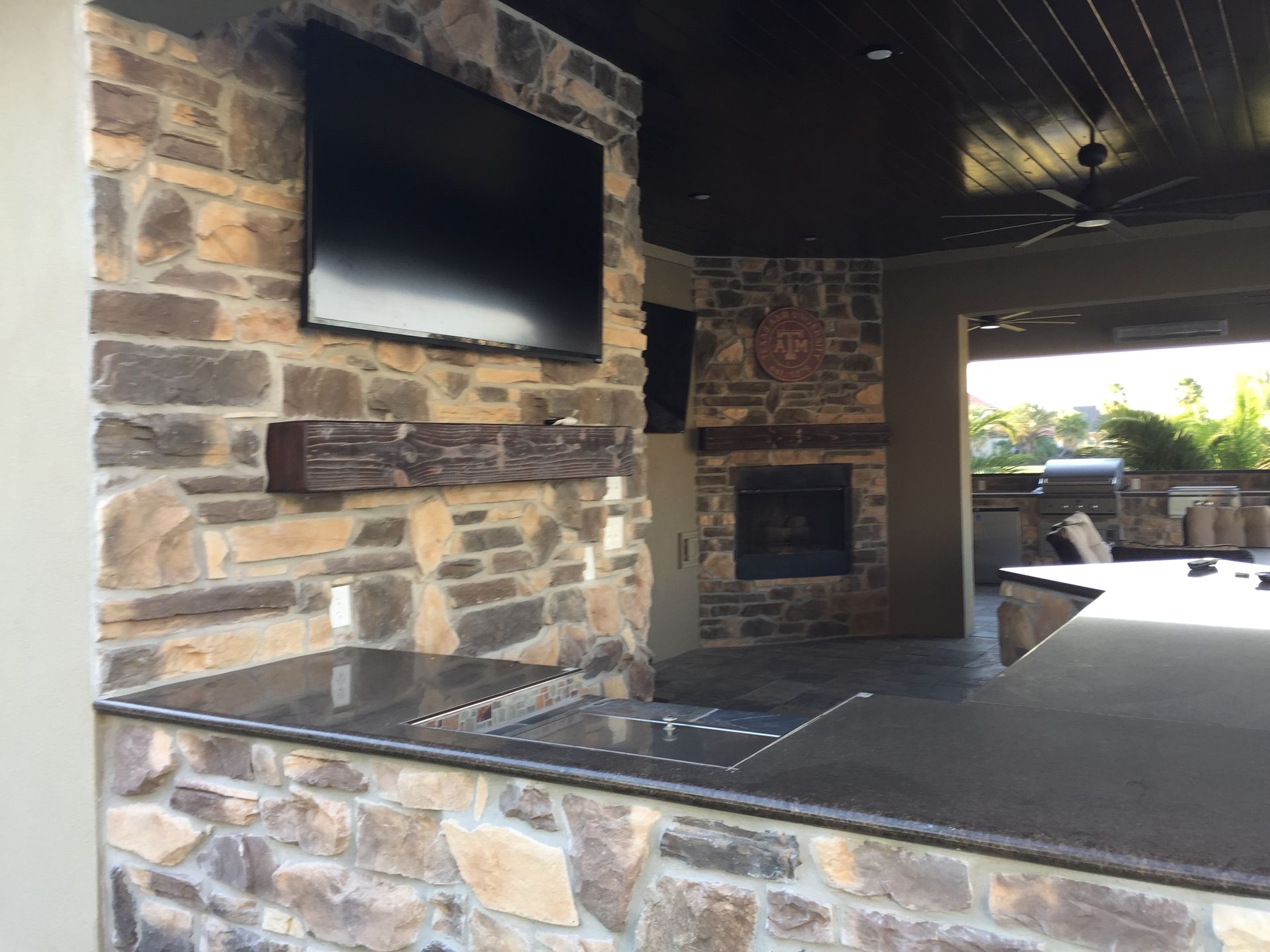 Covered outdoor patio with stone fireplace, mounted TV, and dark ceiling, opening to a bright seating area