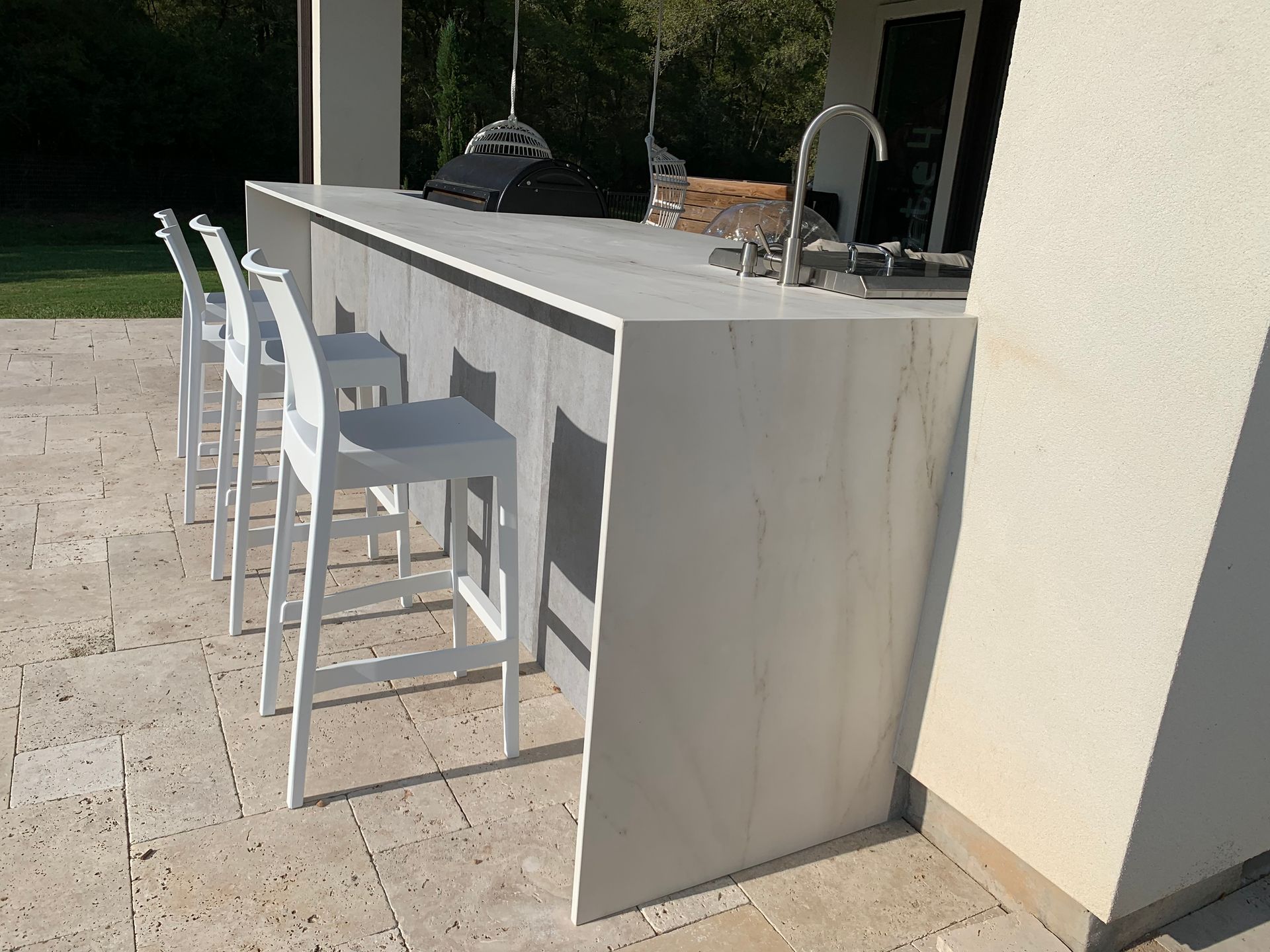 Outdoor patio bar with white stools beside a countertop and sink near a house wall