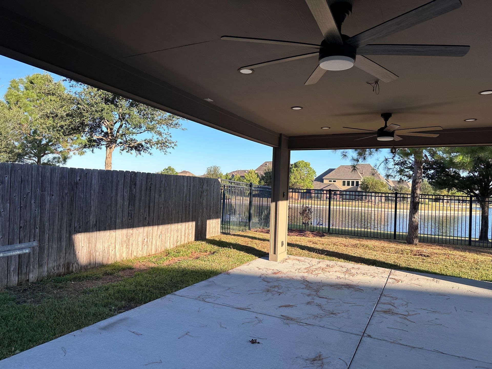 Covered patio with ceiling fans, concrete floor, fence, and grassy backyard beyond the posts.