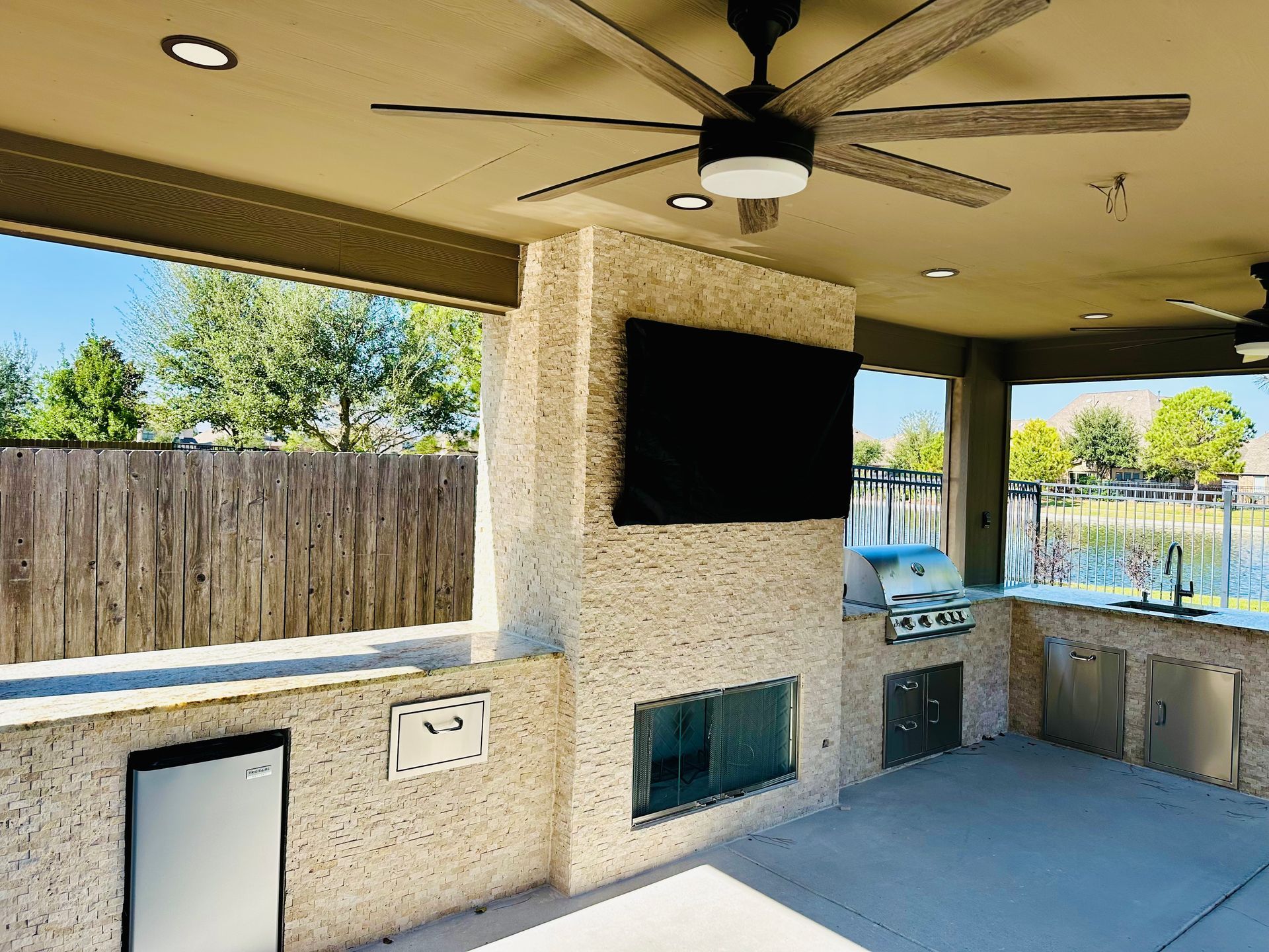 Covered outdoor kitchen with ceiling fans, stone countertops, built-in grill, and a wall-mounted TV beside a pool.