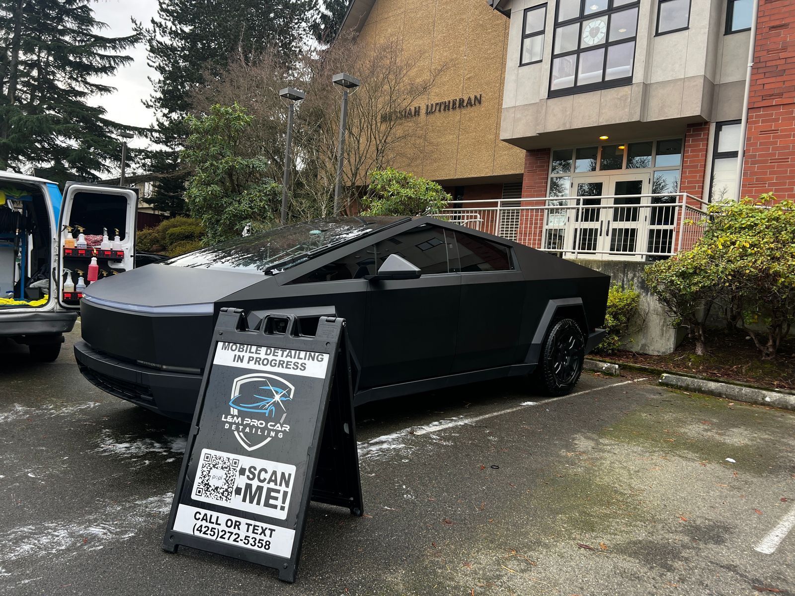 Black Cybertruck parked beside a sandwich-board sign outside a building in a wet lot