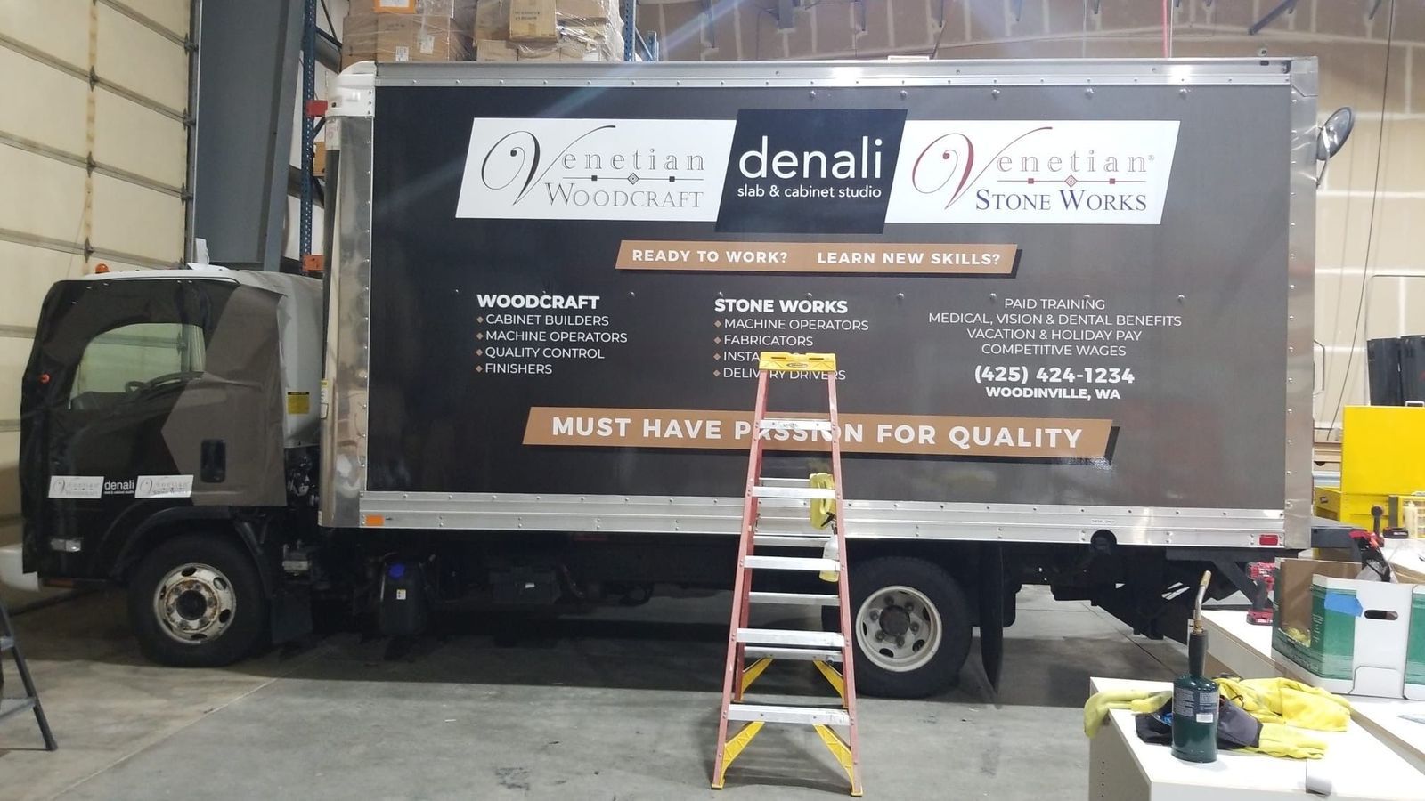 Box truck in a warehouse with a large advertisement banner and a ladder leaning against it.