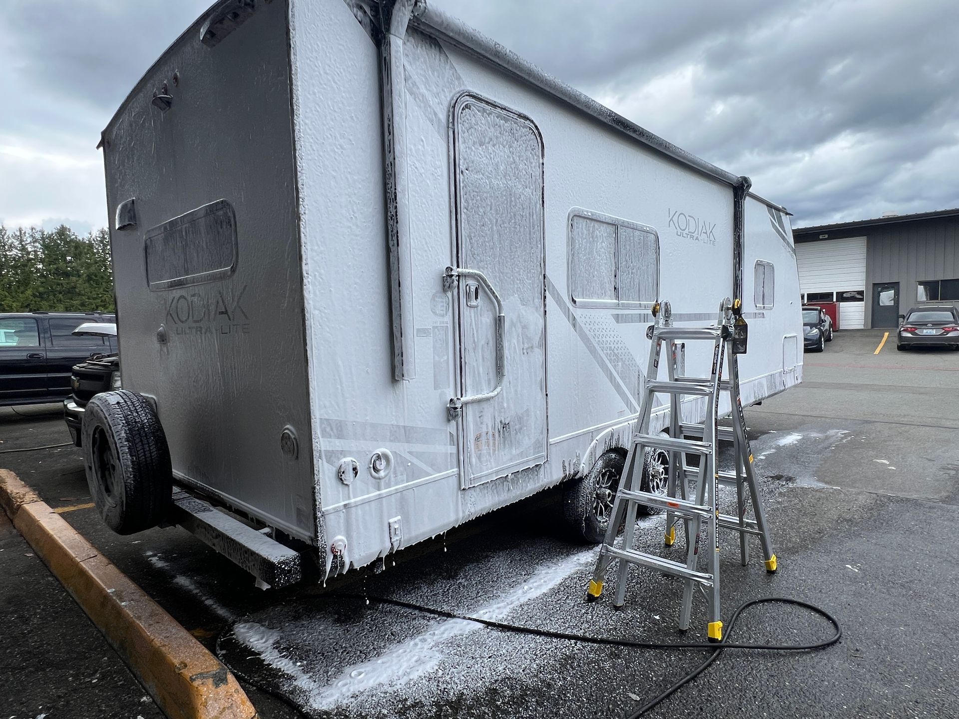 White trailer being washed with a ladder and hose in an outdoor lot under cloudy skies.