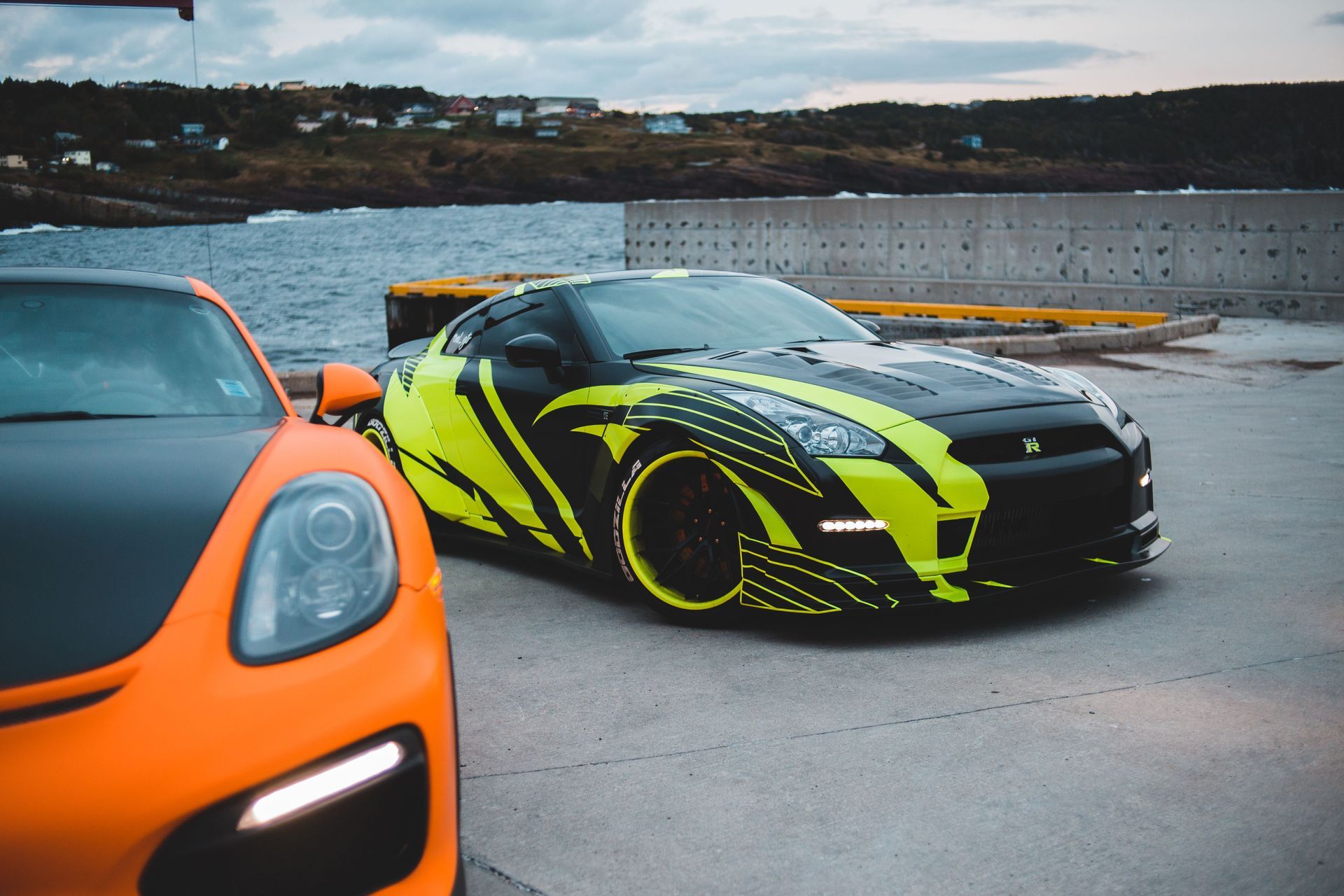 Two sports cars parked on a waterfront road, one orange and one black with neon yellow graphics.