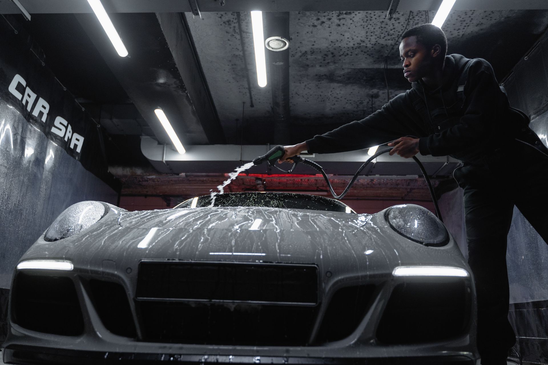 Person rinses a gray sports car in a dim car wash with overhead lights.