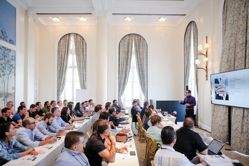 A group of people are sitting at long tables in a conference room.