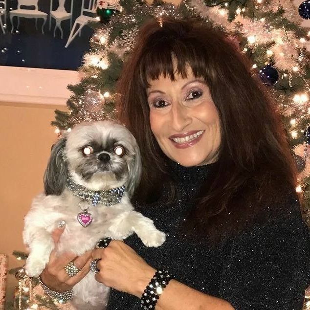 Woman with brown hair smiles, holding a small dog with jeweled collar in front of Christmas tree.