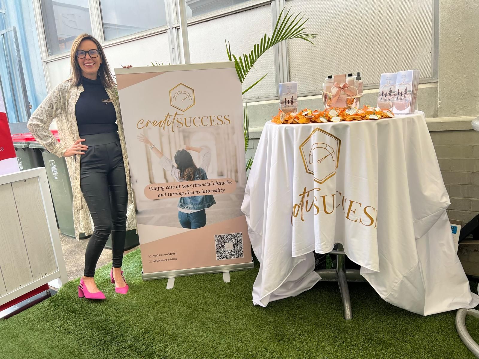 Woman poses by a table displaying branded products. The products include candles and a sign with a logo and text on it.