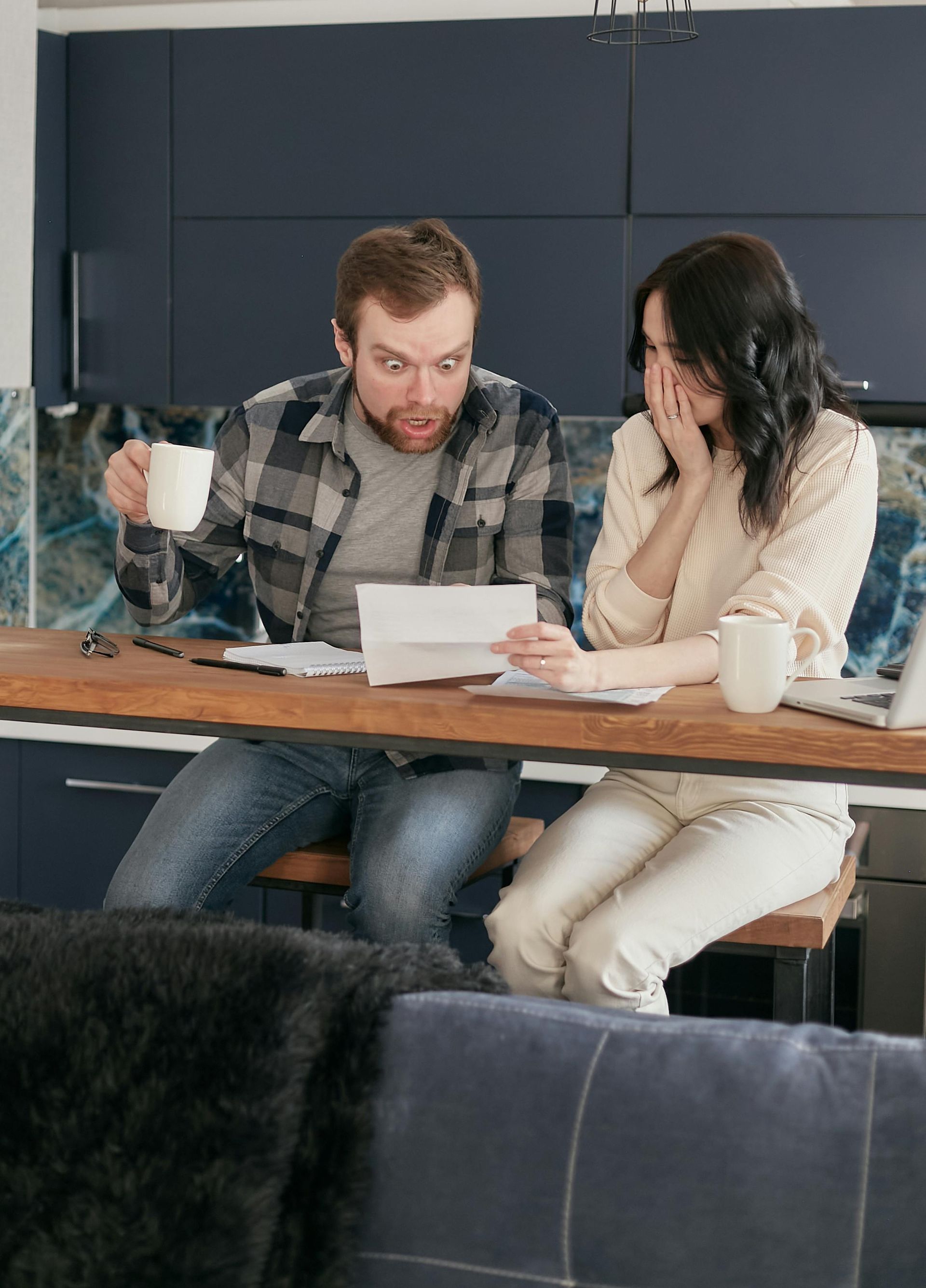 Couple reacts with surprise while looking at paperwork, kitchen setting.