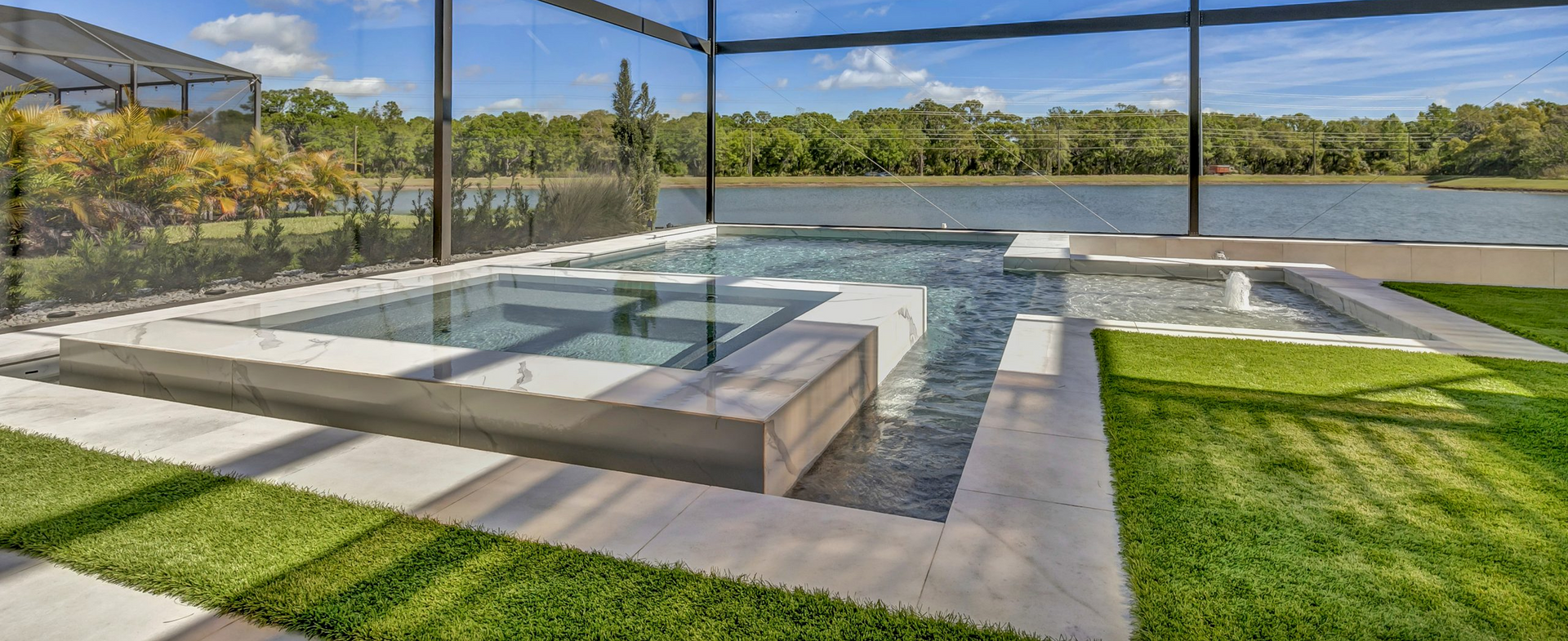 A modern backyard with a pool overlooking a lake, bordered by green grass and concrete paving.