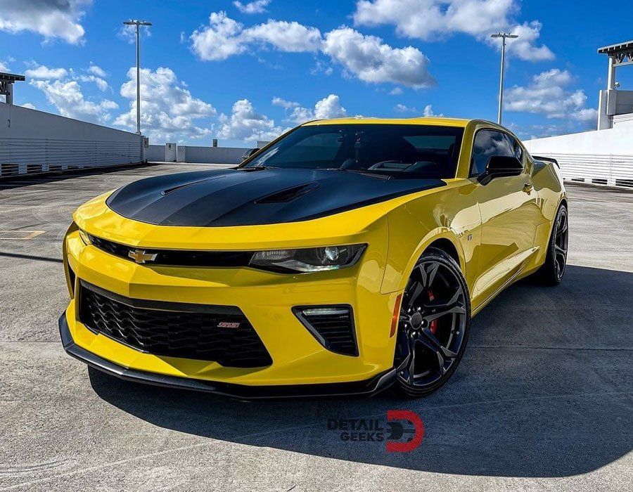 A yellow chevrolet camaro with a black hood is parked in a parking lot.