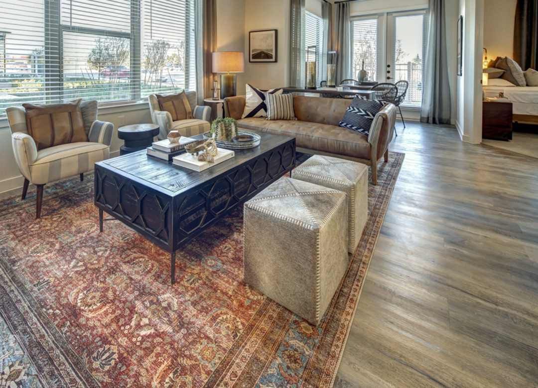 Living room with a brown sofa, patterned rug, and two ottomans. Large windows let in light.
