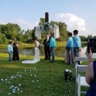 Outdoor wedding ceremony by a lakeside with bride, groom, officiant, and guests on a grassy lawn.