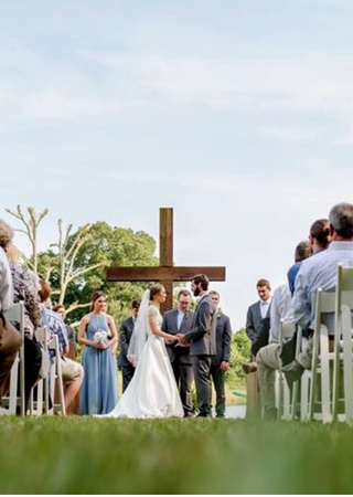 Outdoor wedding ceremony with couple before a wooden cross, guests seated on both sides, bride in white gown