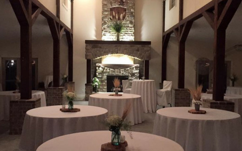 Elegant lobby with round tables draped in white cloths and tall dark beams framing a fireplace.