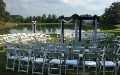 Outdoor wedding setup with white chairs facing a black-draped arch by a lakeside lawn