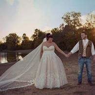 Bride and groom holding hands outdoors at sunset, with long veil trailing behind her.