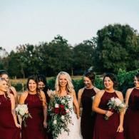 Bride in white dress with bridesmaids in burgundy dresses, holding bouquets outdoors at a wedding