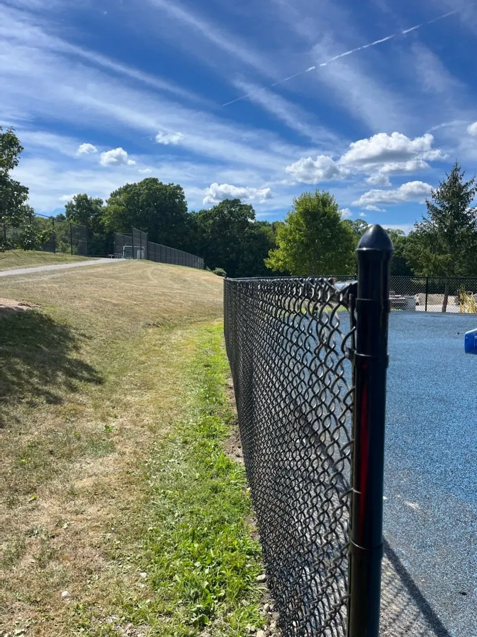 Chain-link fence beside a gravel area and grassy hill under a blue sky with clouds