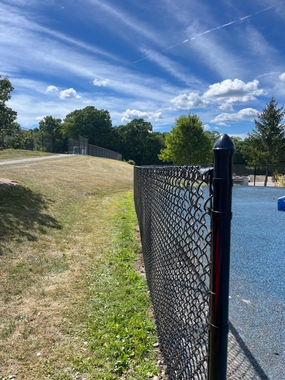Chain-link fence beside a gravel lot and grassy hill under a blue sky with wispy clouds
