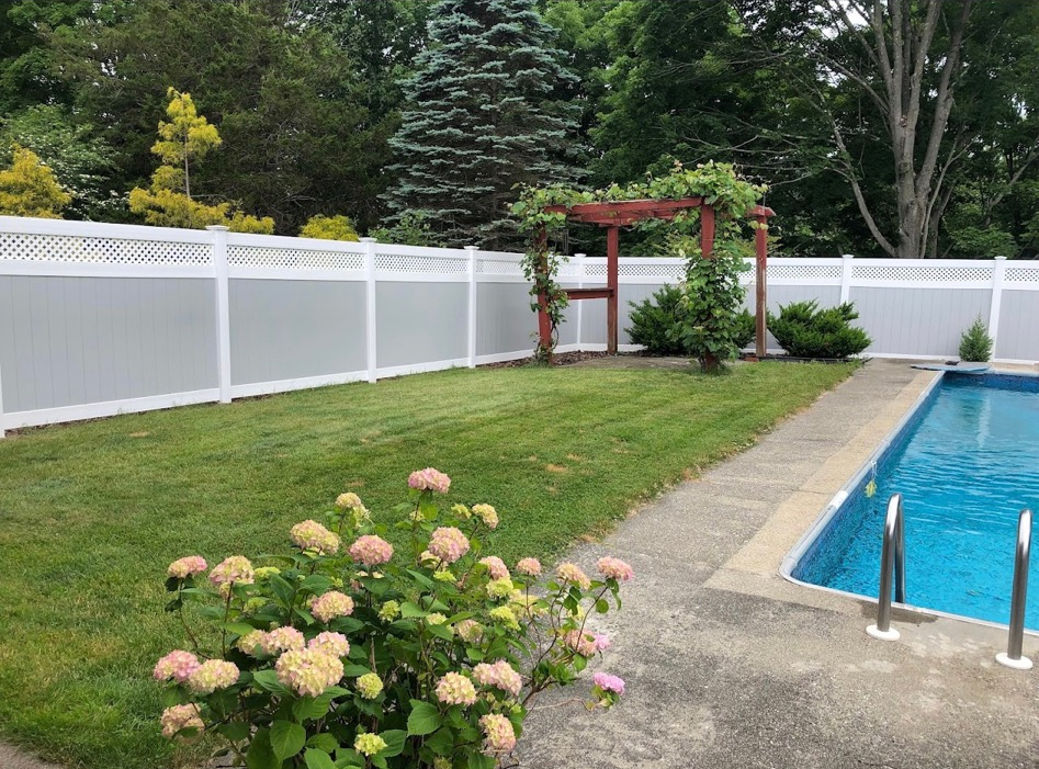 Backyard pool beside lawn, white fence, flowering shrubs, and a wooden pergola surrounded by trees