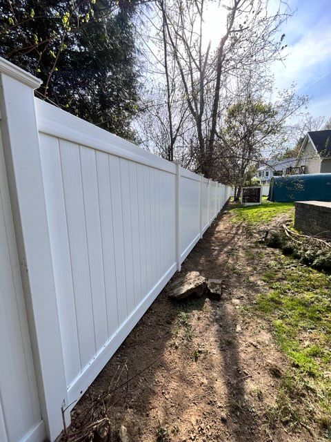 White vinyl fence along a narrow dirt path beside a yard, with trees and a house in the background